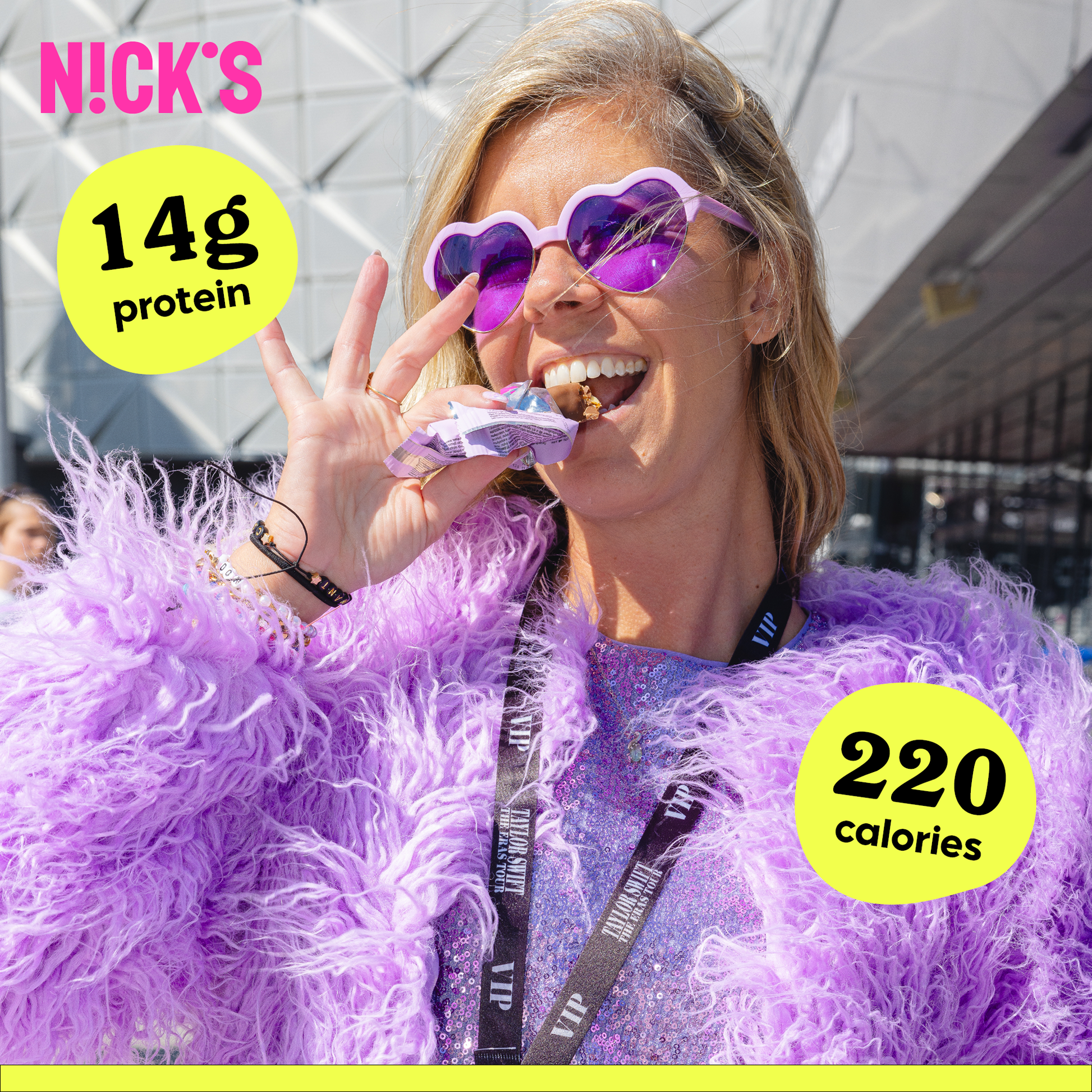 Smiling woman wearing purple faux fur and heart-shaped sunglasses, eating a N!CK'S Almond Chocolate Protein Bar. Text highlights '14g protein' and '220 calories'.