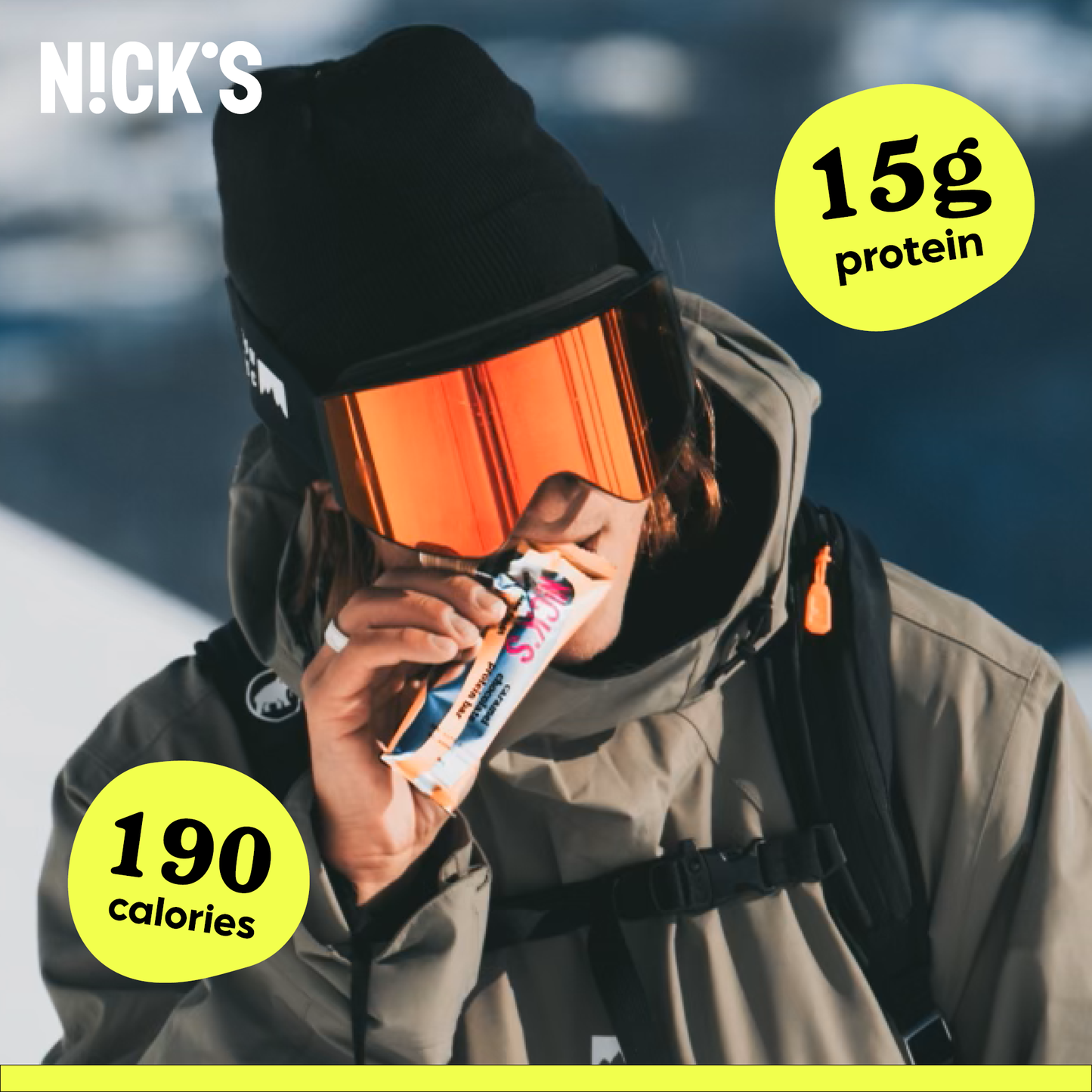 Person wearing winter gear (beanie and goggles) taking a bite of a N!CK'S Caramel Chocolate Protein Bar in a snowy, outdoor setting. Text highlights '15g protein' and '190 calories'.