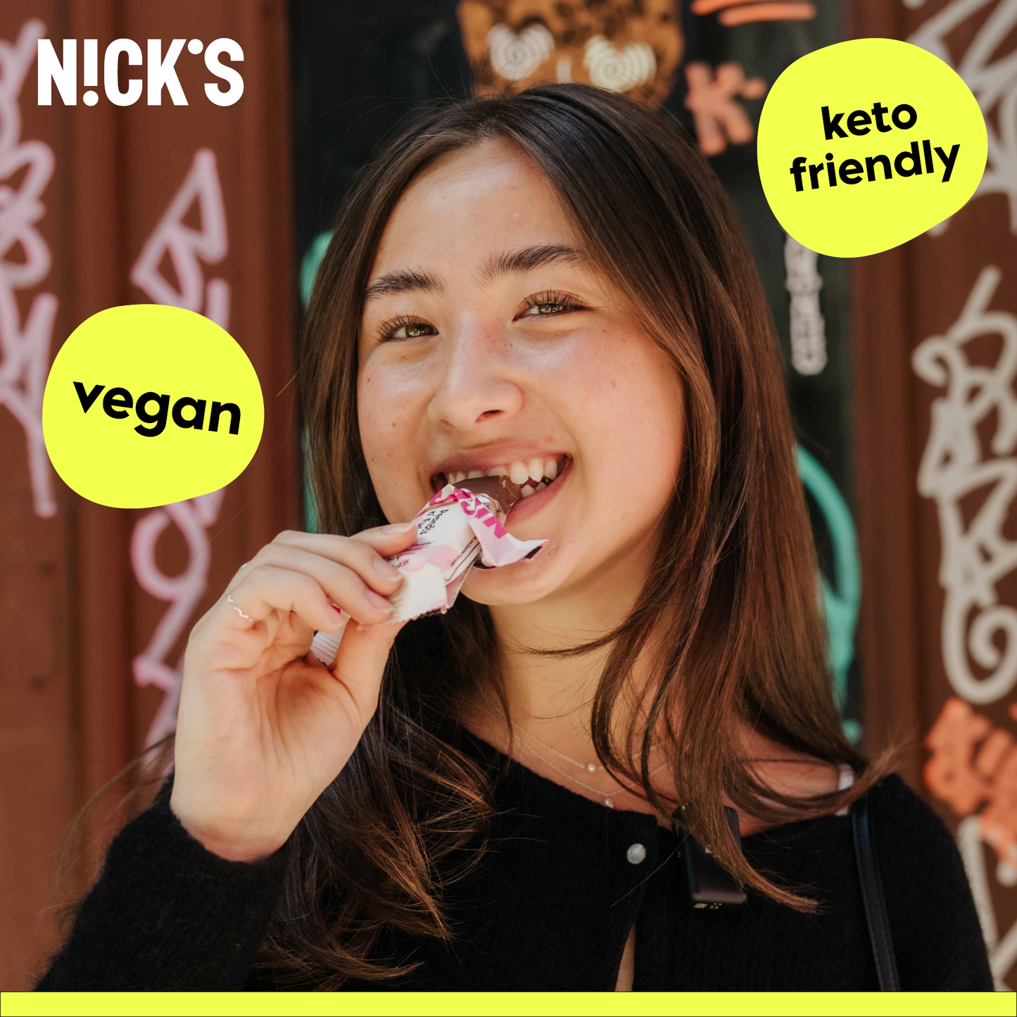 A smiling young woman with dark hair stands in front of a graffiti-covered wall, taking a bite from an unwrapped Cherry Crunch N!CK'S bar. Yellow text bubbles overlaid on the image advertise the product as "vegan" and "keto friendly".