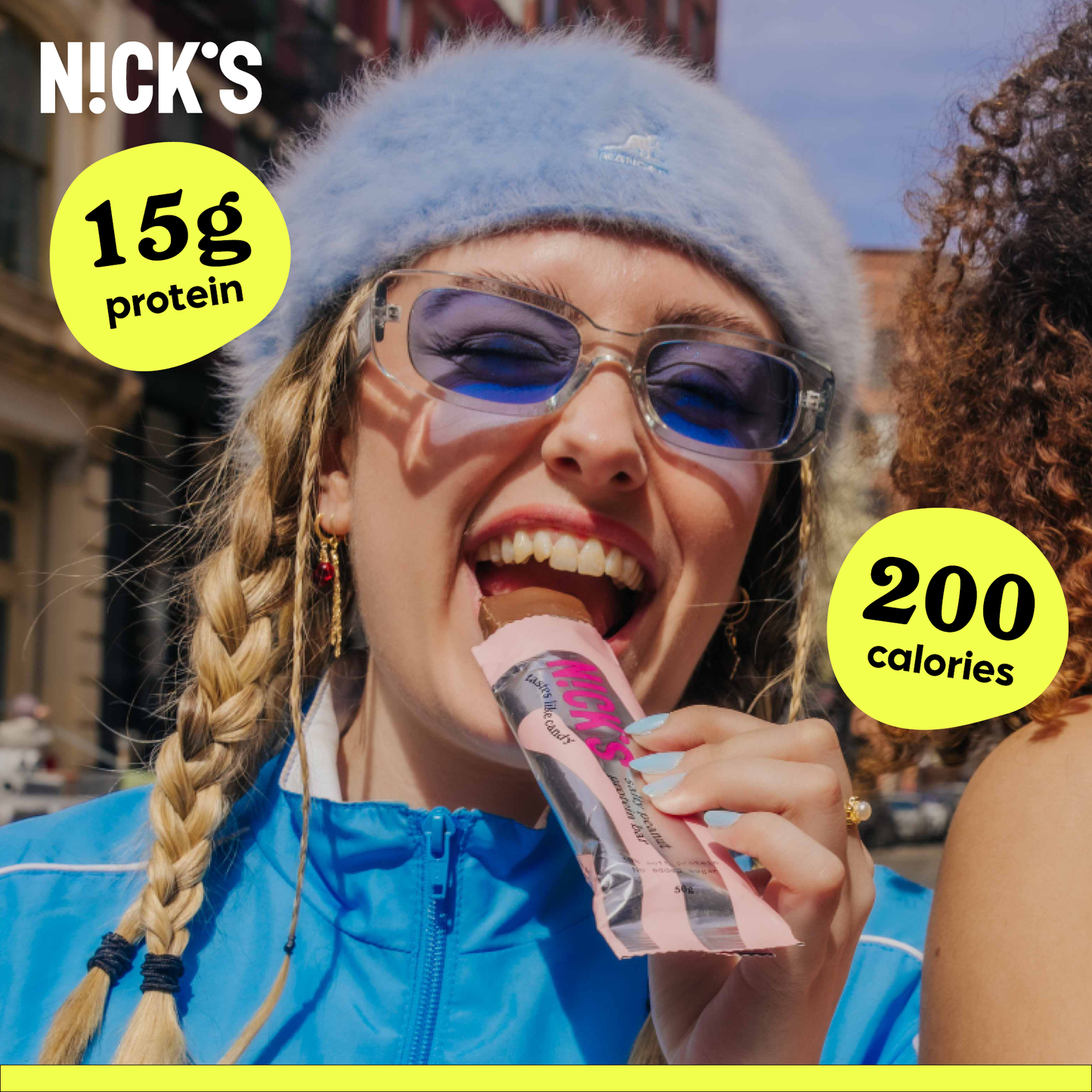 Smiling young woman wearing a blue fuzzy hat and sunglasses, taking a bite of a N!CK'S Salty Peanut Protein Bar. Text highlights '15g protein' and '200 calories'.